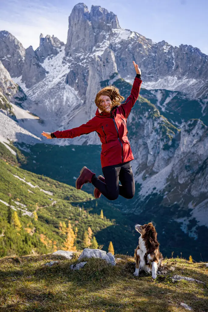 Frau springt in die Luft, Hund schaut zu ihr hoch. In den Bergen Österreichs